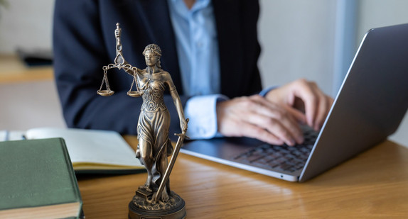 Anwalt im dunklen Anzug tippt auf einem silbernen Laptop. Eine bronzene Justitia-Figur steht auf dem Schreibtisch aus Holz. Ein aufgeschlagenes Notizbuch und ein Stift liegen daneben. Moderne digitale Anwaltskanzlei mit dem traditionellen Symbol der Justiz im Büro.