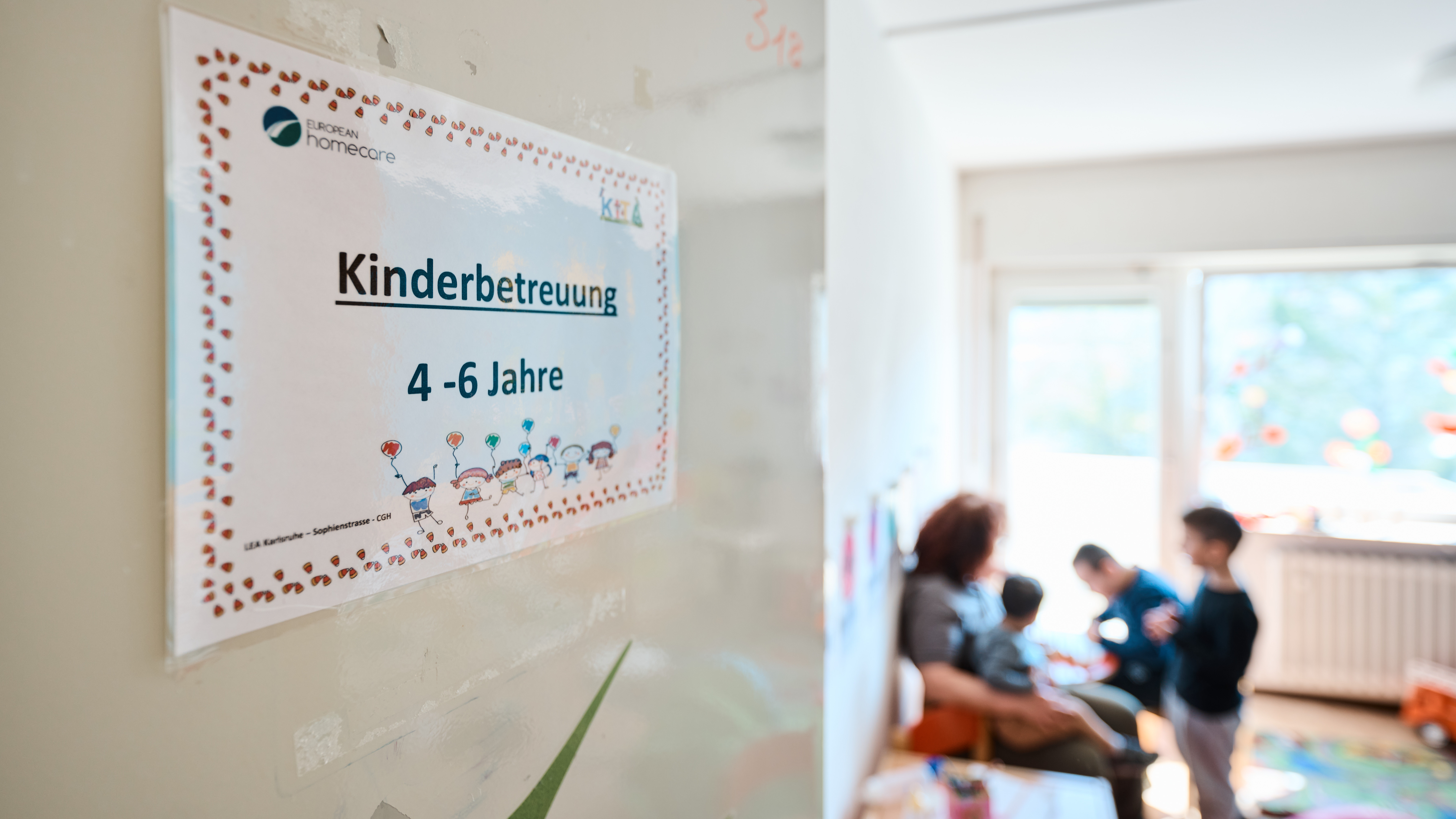 Das Bild zeigt den Blick in einen Raum, in dem eine Betreuerin sich mit Kindern beschäftigt. An der Tür hängt ein Schild mit der Aufschrift: Kinderbetreuung 4 bis 6 Jahre