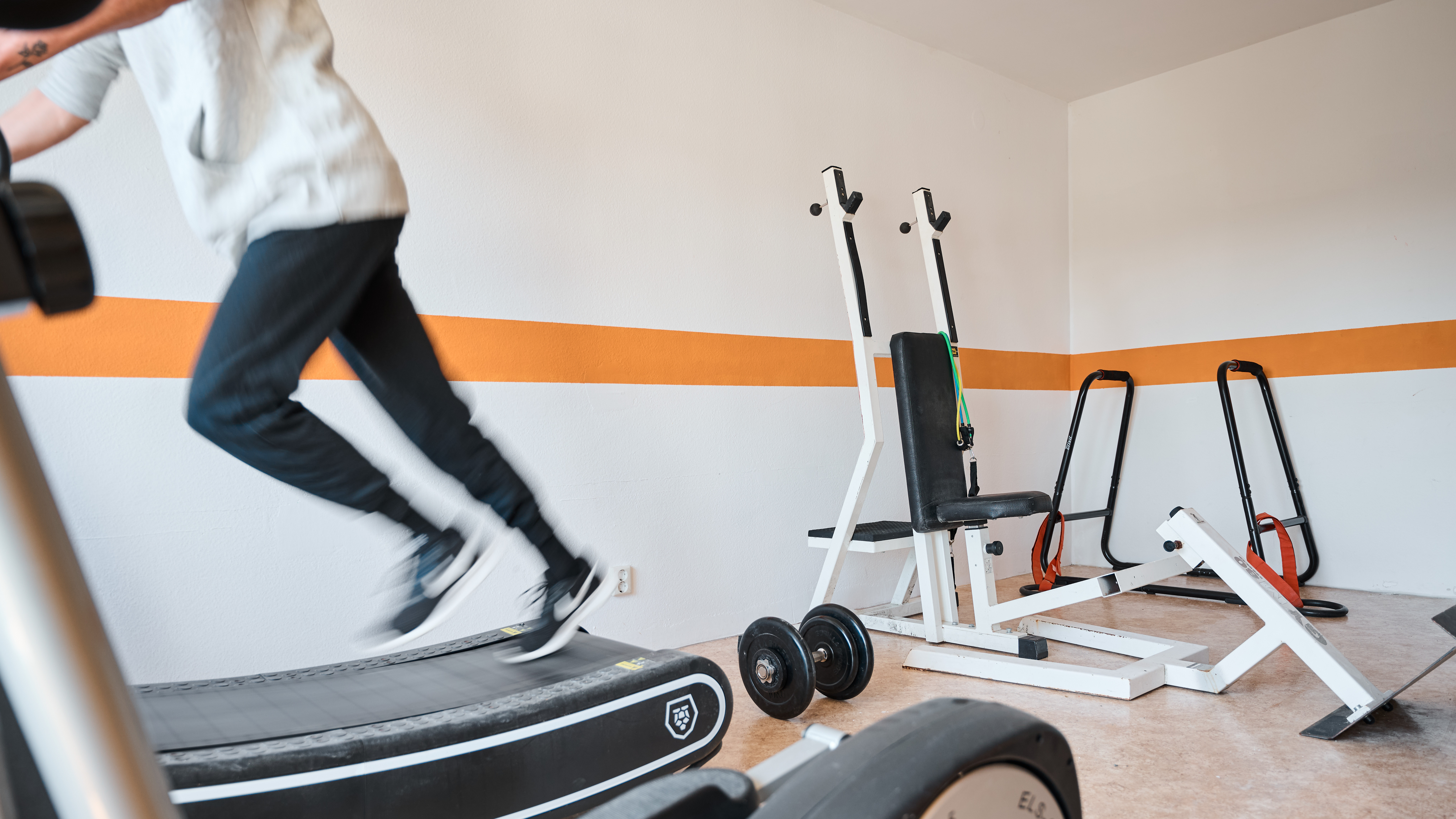 Das Bild zeigt eine Person auf dem Laufband in einem Raum mit verschiedenen Fitnessgeräten.