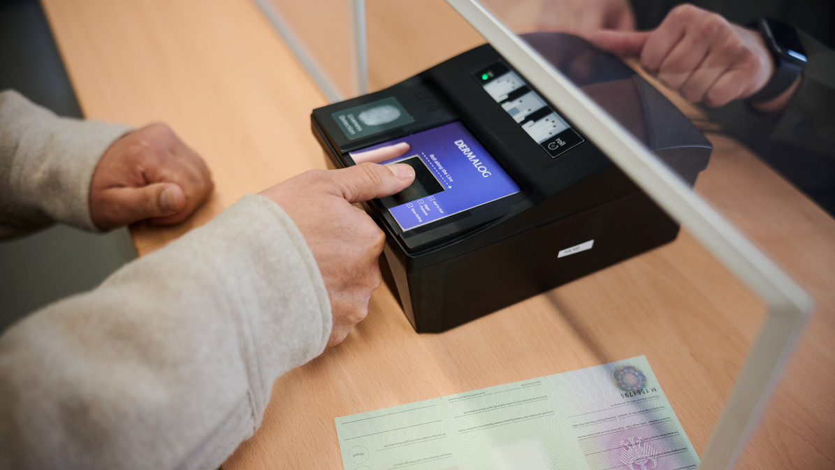 Das Bild zeigt, wie bei einer Person ein Fingerabdruck mit Hilfe eines Fingerabdruckscanners genommen wird. 